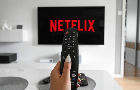 Eine Hand hält eine Fernbedienung, im Hintergrund ein TV mit Netflix-Logo 