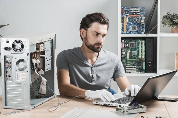 Mann sitzt an einem Laptop, neben sich ein geöffnetes PC-Gehäuse 