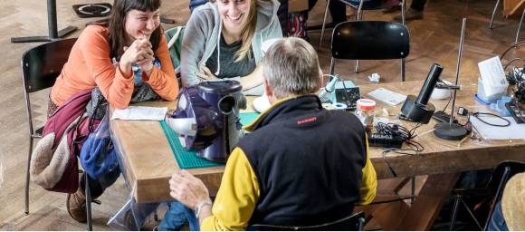 3 Personen sitzen um einen Tisch mit Elektrogeräten drauf 