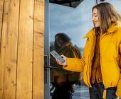 Eine Frau in gelber Windjacke hält ihr Smartphone ans Schloss einer Haustür