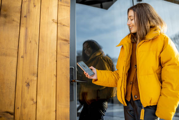 Eine Frau in gelber Windjacke hält ihr Smartphone ans Schloss einer Haustür 