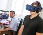 Ein Mann trägt eine Virtual-Reality-Brille, im Hintergrund sitzt ein zweiter Mann an einem Computer an einem Tisch