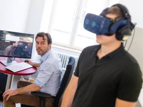 Ein Mann trägt eine Virtual-Reality-Brille, im Hintergrund sitzt ein zweiter Mann an einem Computer an einem Tisch 