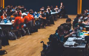 In einer Halle sitzen zahlreiche junge Leute mit ihren Notebooks an Tischen 