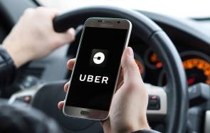 Blick über die Schulter einer Person, die im Auto am Steuer sitzt und ein Smartphone hält, das ein Uber-Logo zeigt 