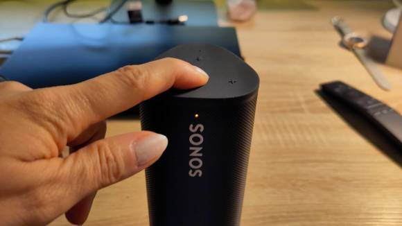 Ein Finger drückt auf der Oberseite des Sonos Roam einen Knopf, eine orange LED leuchtet