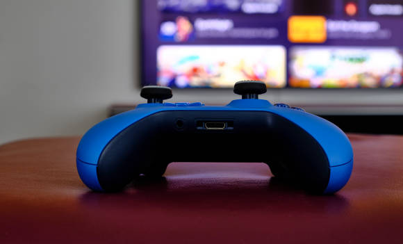 Auf einer Unterlage aus rotem Leder liegt ein blauer Xbox-Controller; im Hintergrund ist unscharf die Oberfläche von Apple TV zu sehen