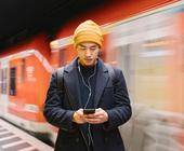 Ein junger Mann steht an einem Bahngleis und schaut auf sein Smartphone