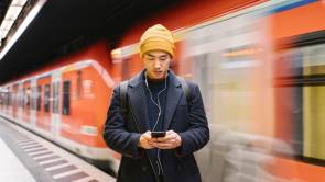Ein junger Mann steht an einem Bahngleis und schaut auf sein Smartphone 