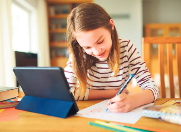 Symbolbild zeigt ein Mädchen, das neben einem Tablet sitzt und auf ein Blatt Papier kritzelt 