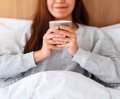 Eine Frau im grauen Pullover sitzt im Bett und trinkt aus einer Tasse