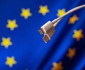 Symbolbild zeigt einen Lightning- und einen USB-C-Stecker vor einer Europa-Flagge