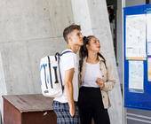 2 junge Menschen am Bahnhof vor Reiseinfowand