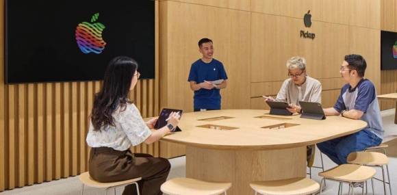 Symbolbild zeigt vier Personen, die mit ihren Tablets an einem Tisch in einem Apple-Shop sitzen oder stehen 